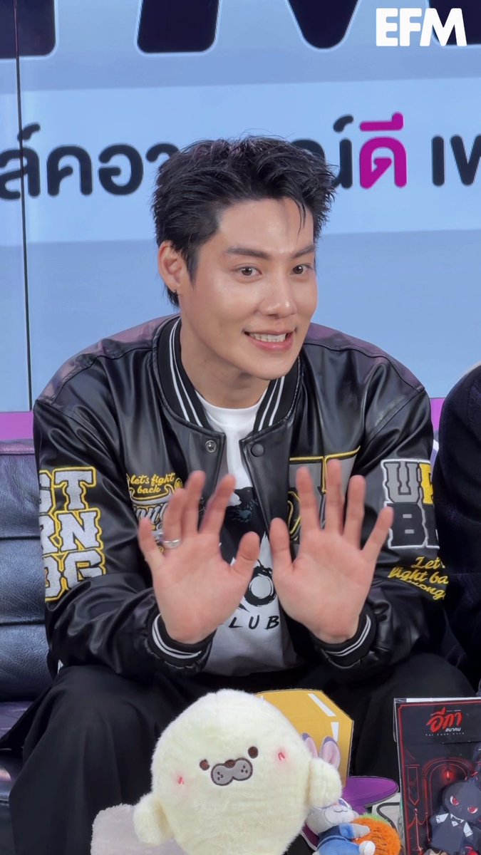 An EFM-branded photo of a smiling young man in a black varsity jacket waving both hands, seated next to plush toys in front of a Thai-language backdrop.