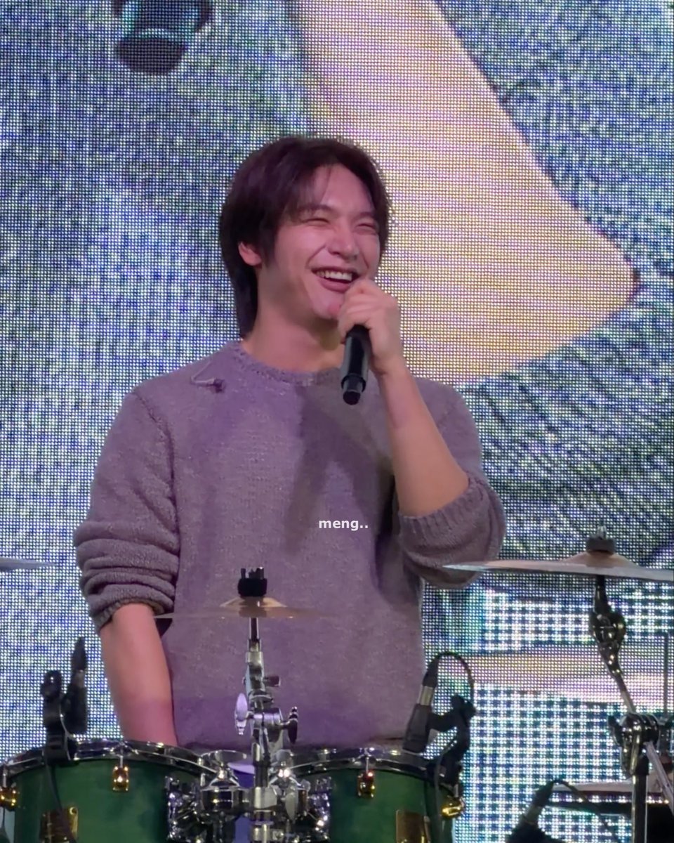 A smiling man with dark hair in a gray sweater holding a microphone while standing behind a drum set on a stage; the text 'meng..' is visible.