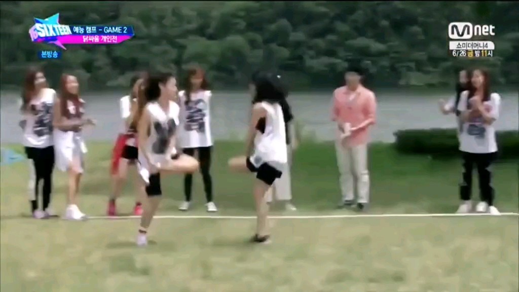 This image appears to be a screenshot from the Mnet show "SIXTEEN," as shown in the top left corner, which was the survival show that formed the K-pop group TWICE. The overlay text indicates this is from the "예능 캠프 - GAME 2" (Entertainment Camp - Game 2) round, specifically the "왕좌를 거머쥐면" (Seize the Throne) challenge.

In the image, a chaotic outdoor game scene unfolds on a grassy field, where several contestants wearing white tops and black shorts/pants are engaged in what looks like a competitive elimination game. The players are seen in dynamic poses—some appearing to push or knock each other down in an attempt to claim a central throne or position. The caption references how Chaeyoung aggressively knocked down her friend Somi during this intense round, prioritizing victory over their friendship.