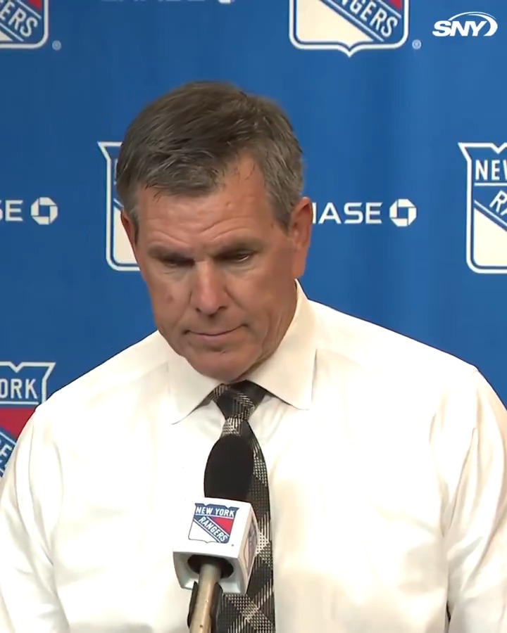 Mike Sullivan, head coach of the New York Rangers, is shown at a press conference with his head bowed, speaking into a microphone branded with the Rangers logo. His downcast expression suggests he is reflecting on Igor Shesterkin's historic goal, as the post quotes him calling it "a great moment" that highlighted the goalie's puck-handling strength.
