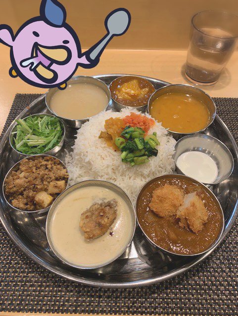 An Indian thali meal served on a metal platter featuring rice, dal, curries, vegetables, and a fried item, with a glass of water and a cartoon sticker overlay in the corner.
