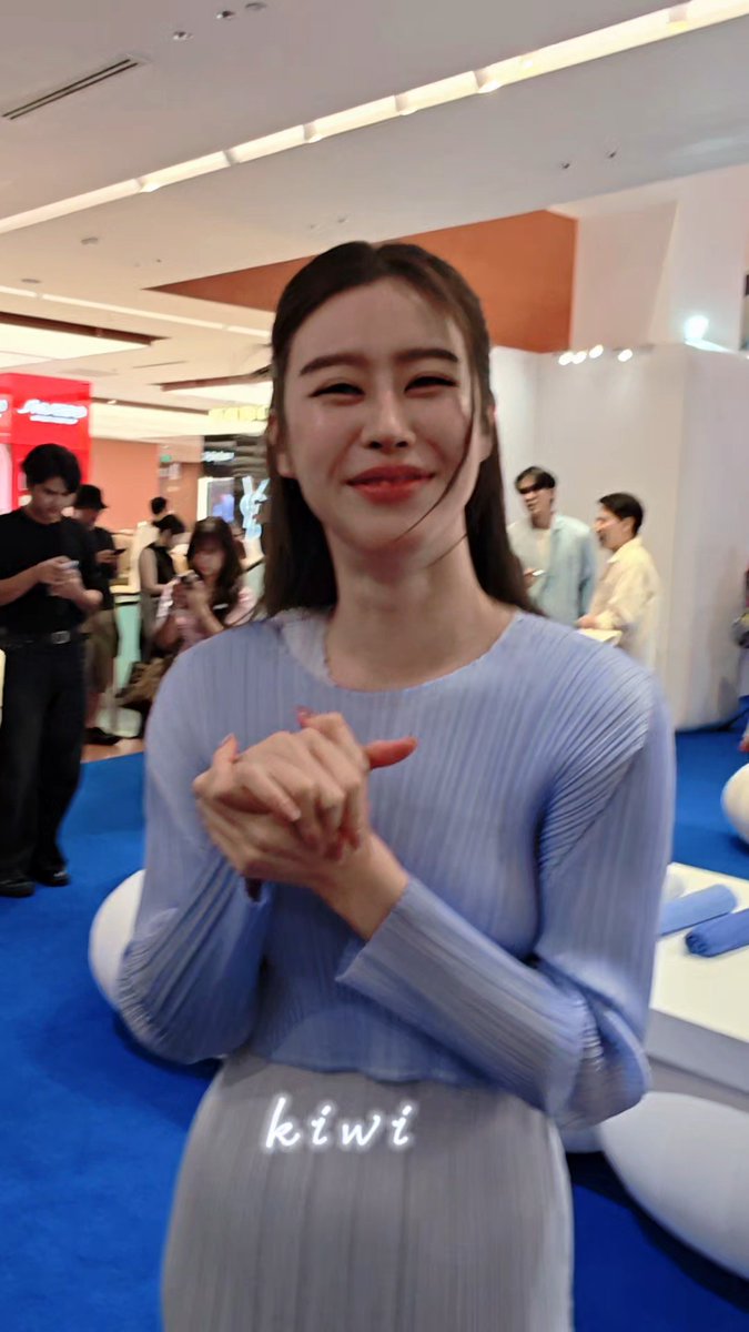A young woman with long brown hair smiles at the camera wearing a light blue long-sleeved ribbed top and matching pants, with the text 'kiwi' overlaid on the image, standing at what appears to be an exhibition or trade show.