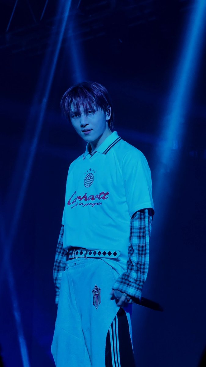 A young male performer stands on stage under blue lighting, wearing a white 'Carhartt trust in progress' polo layered over a plaid shirt, holding a microphone.