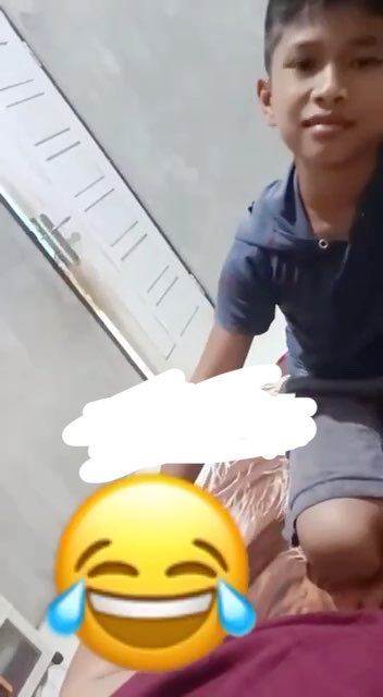 A child sits on a bed in a selfie-style video with a laughing-crying emoji covering part of the scene, shared via tribunvideo.com. The short clip appears to be a candid, lighthearted moment uploaded to the video platform.