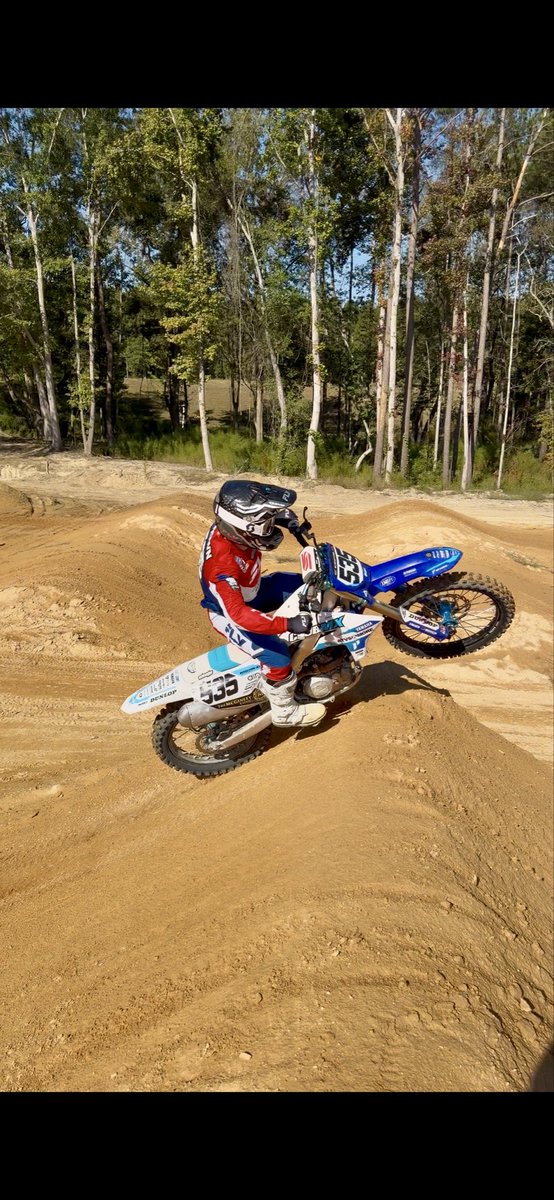 A motocross rider wearing a red, white, and blue jersey and helmet with number 335 navigates a dirt track, performing a wheelie on a blue Yamaha dirt bike.