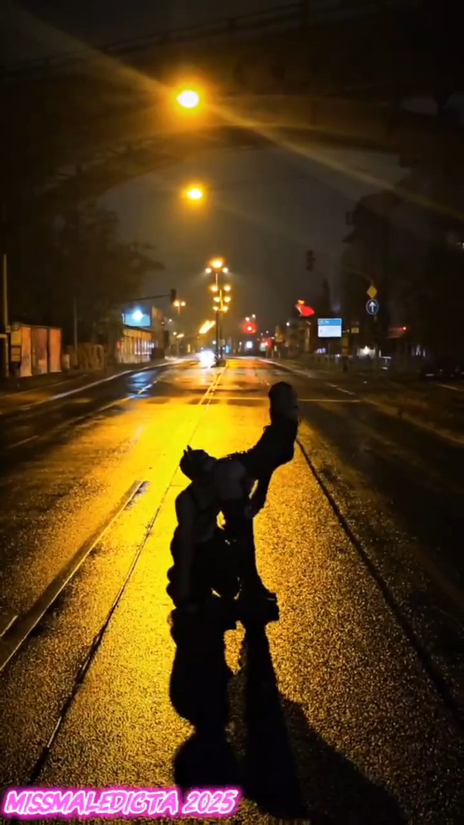 A nighttime photograph of an empty, wet city street illuminated by amber streetlights, featuring a silhouetted statue in the foreground and the watermark 'MISSMALEDICTA 2023' in pink text.