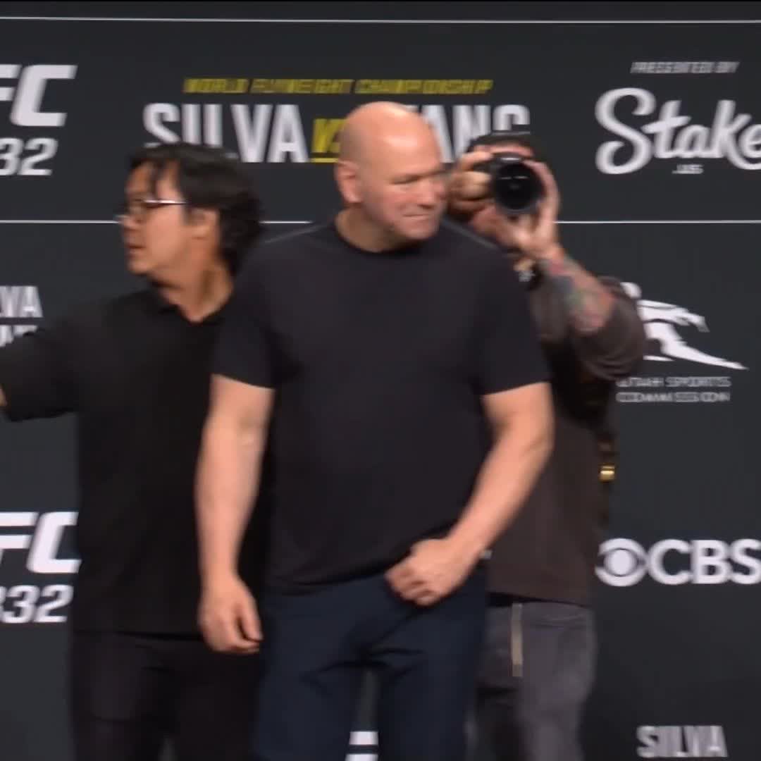 Two men, including a bald fighter in a black t-shirt, stand on stage at a UFC 332 press event for the co-main fight between Deiveson Figueiredo and Payton Talbott, with a photographer capturing the moment against a backdrop featuring UFC 332, Stake.us, and CBS logos.