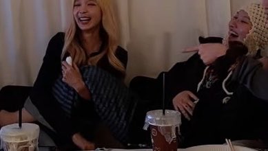 Two women are laughing heartily while sitting on a couch, holding iced drinks and a pillow, one pointing playfully at the other.