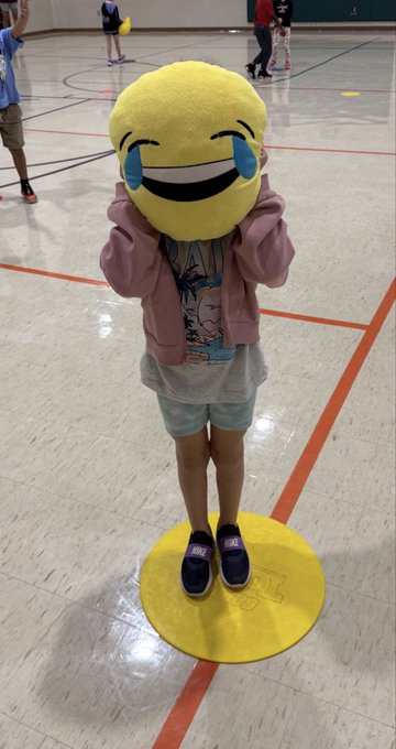 A child standing on a yellow floor mat in a gymnasium, holding a large yellow emoji plush pillow with laughing-crying tears covering their face.