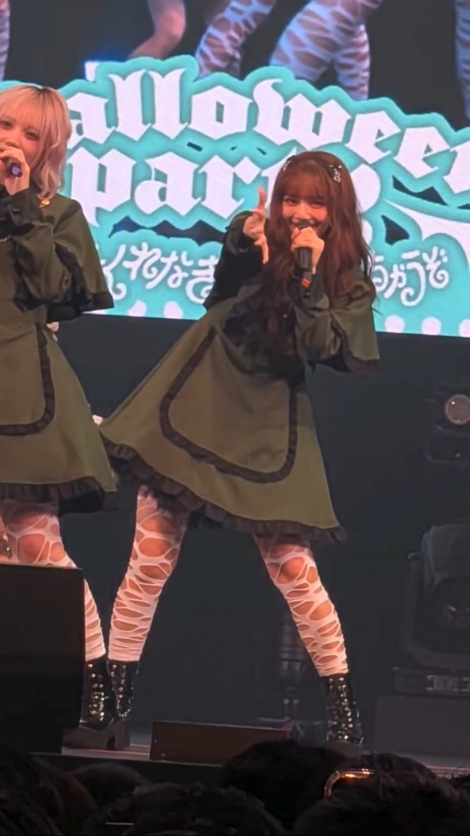 Two women perform on stage, one with brown hair holding a microphone and pointing while wearing an olive green dress over patterned tights, with a banner reading 'aMonster Party' behind them.