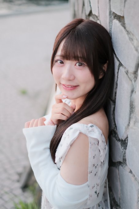 A smiling young woman with long brown hair leans against a stone wall, wearing a white floral-patterned top that exposes her shoulders.