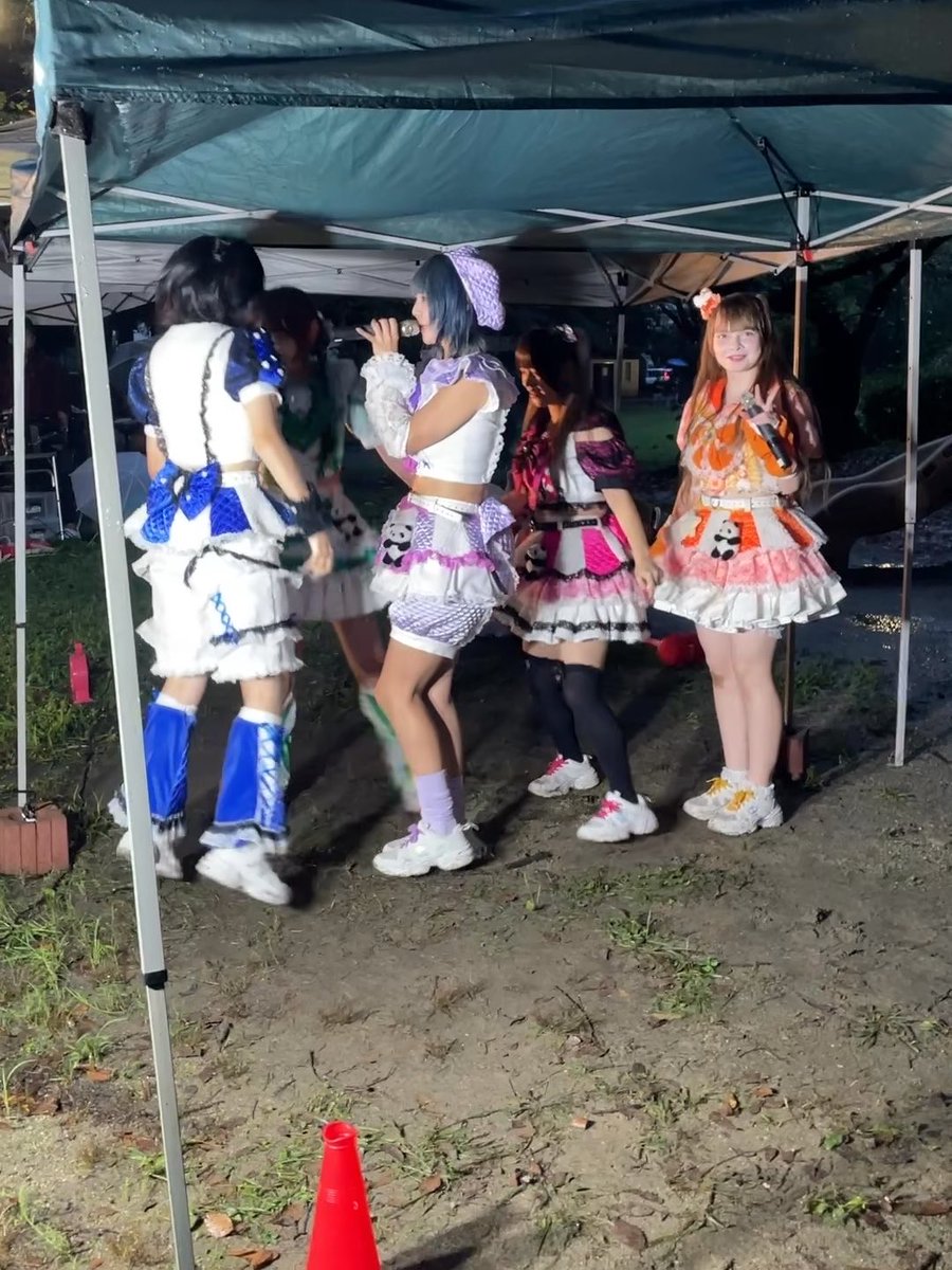 Five young women in elaborate, colorful idol-style costumes perform under a green tent outdoors, with one singing into a microphone.