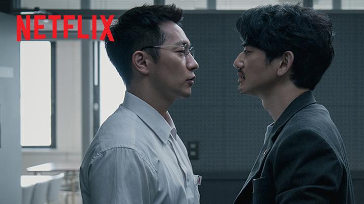 Two men—one wearing wire-frame glasses and a light button-up shirt, the other in a dark blazer with tousled hair—stand face-to-face in a tense, close standoff in what appears to be a stark interior. The "NETFLIX" logo is prominently displayed in red in the upper left corner, tying the image to the streaming platform's hard-boiled crime series featuring Korean detective Son Suk-ku and Japanese detective Nagayama Eita in a cross-border investigation.