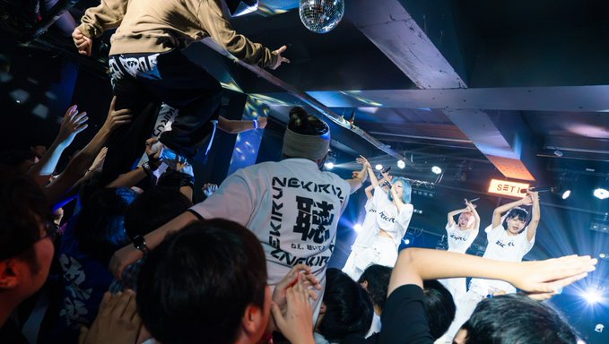 A lively concert scene where a performer is crowd-surfing over an audience, while a group of performers in white outfits stands on stage in the background beneath a disco ball.
