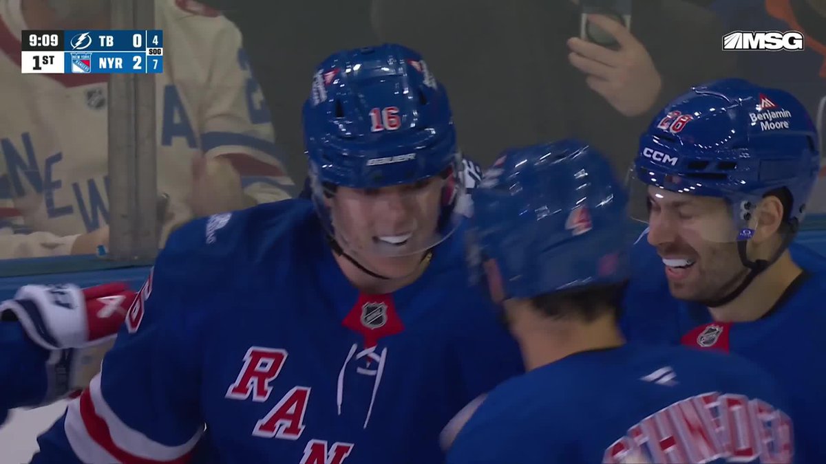New York Rangers players wearing blue jerseys with #16 and #20 helmets celebrate on the ice after a goal, with the scoreboard showing NYR leading TB 2-0 at 9:09 of the 1st period on MSG Network. The image captures the celebration that follows the goal described in the caption, with Dorofeyev likely scoring his first goal as a Ranger off a feed from Bjorkstrand.