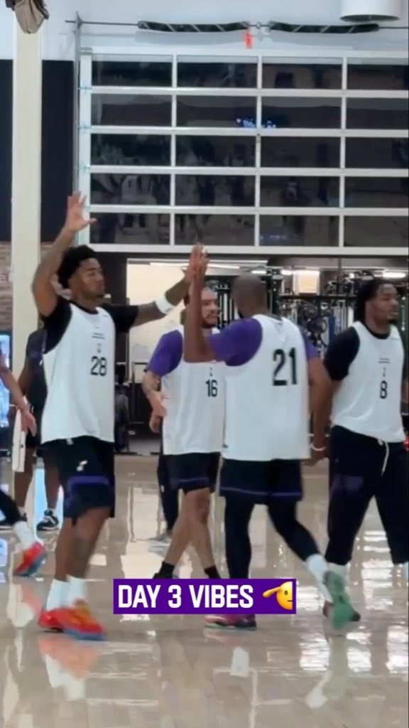 Basketball players wearing numbered practice jerseys (16, 21, 28, and 8) high-five each other during a training session on an indoor court, with a purple banner reading "DAY 3 VIBES" displayed across the image—capturing the team's energetic camaraderie as they put in the work.
