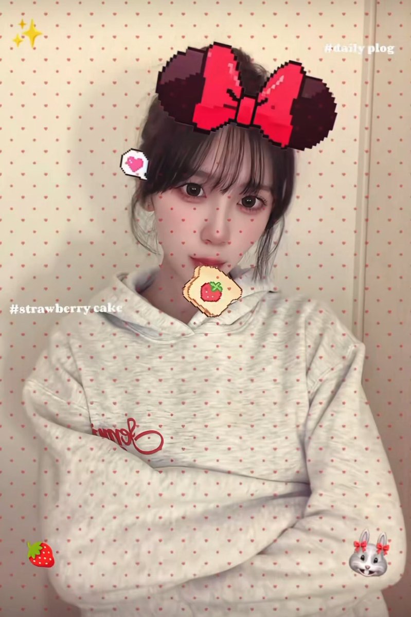 A selfie of a young woman wearing a white hoodie, edited with pixel art stickers including a red bow, a strawberry cake slice, and text overlays '#daily plog' and '#strawberry cake'.