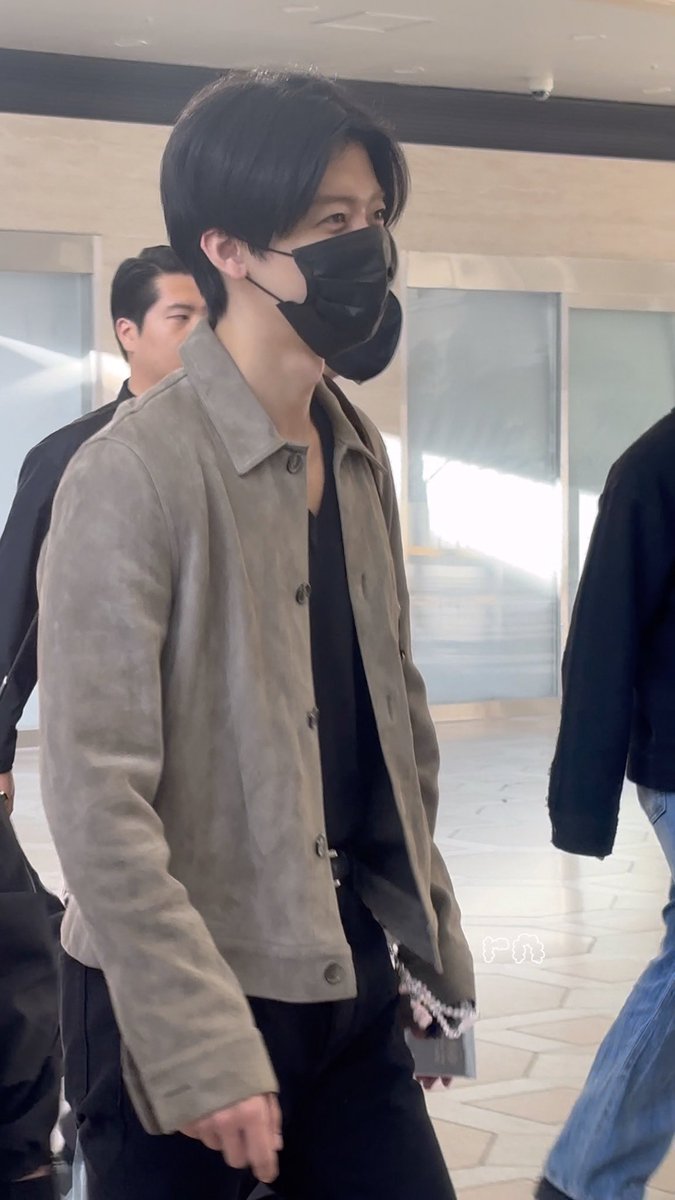 A young man with dark hair wearing a black face mask and a taupe suede jacket walks through what appears to be an airport or public building.