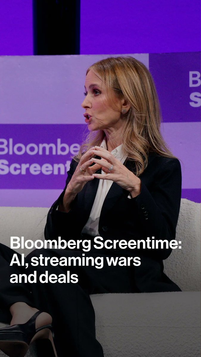 A blonde woman in a black blazer and white blouse speaks with animated hand gestures at the Bloomberg Screentime event, as indicated by the purple branded backdrop and on-screen text reading "Bloomberg Screentime: AI, streaming wars and deals."