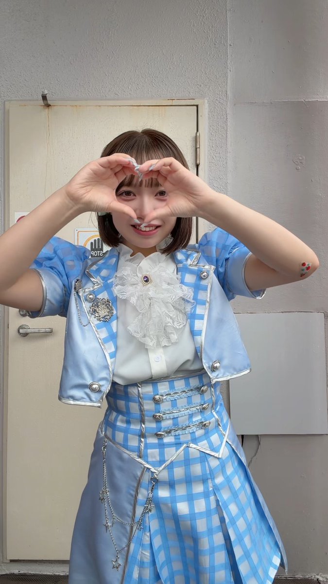 A young woman in a blue and white checkered idol-style outfit with a ruffled jabot makes a heart shape with her hands in front of a plain door.