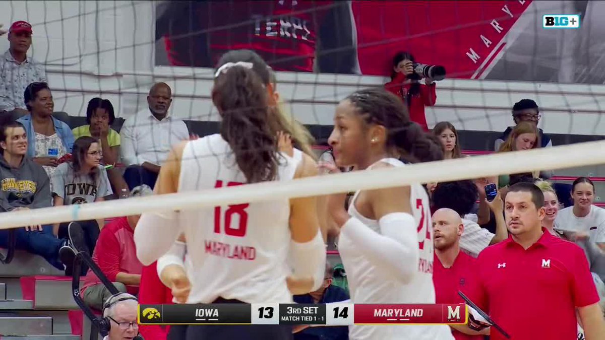 A close-up view through a volleyball net of Maryland players in white jerseys during a game against Iowa, with a scoreboard graphic showing 'IOWA 13' and 'MARYLAND 14' in the 3rd set.