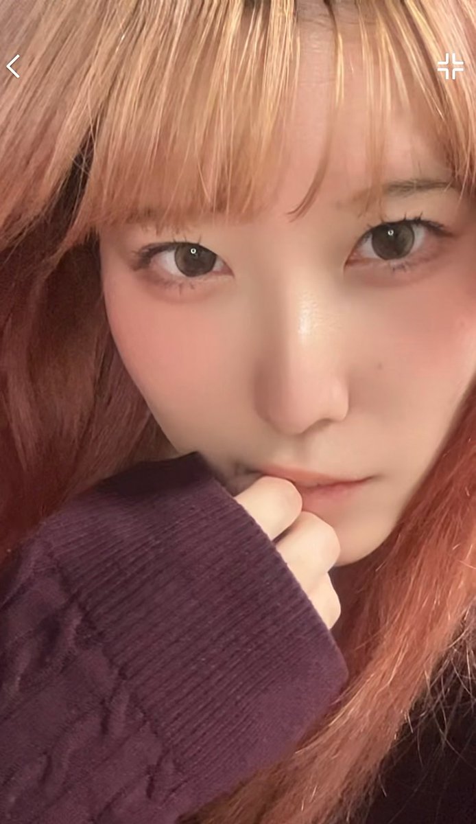 A close-up selfie of a young woman with blonde and pink hair wearing a dark purple sweater, looking directly at the camera.