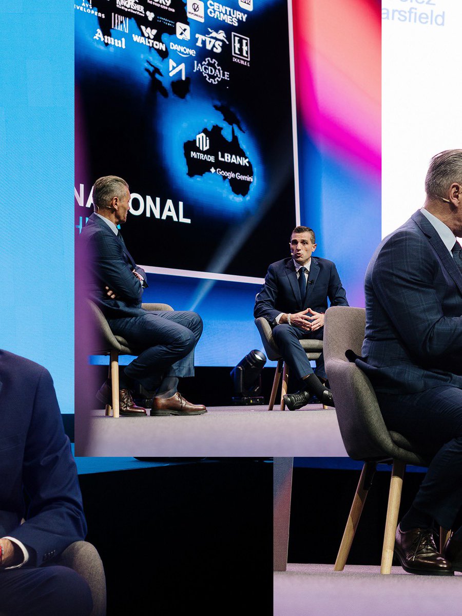 Three men in suits seated on stage in a panel discussion at an event with a backdrop displaying sponsor logos including Mtrade, LBank, Google Gemini, and various corporate brands.