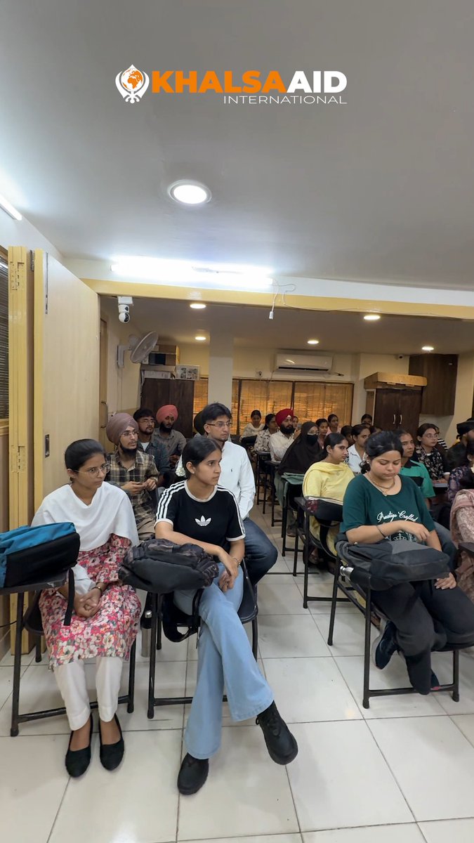 A group of people seated in a classroom or community room under the Khalsa Aid International logo, including a young woman in a black Adidas shirt and white pants in the foreground.