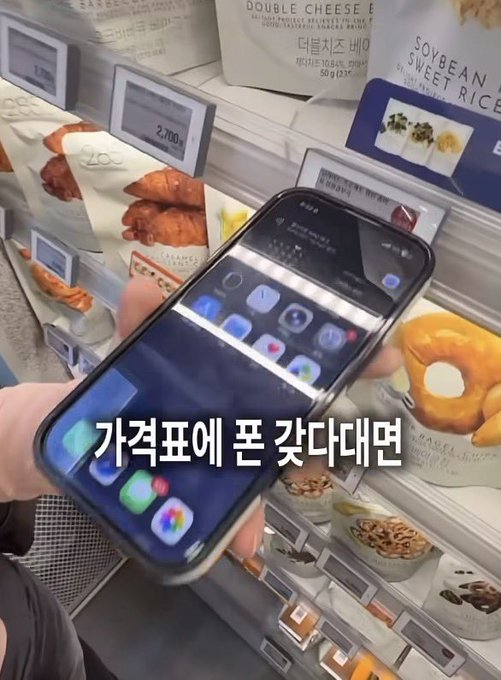 A person holding a smartphone displaying the iOS Control Center and app icons in front of a Korean convenience store shelf stocked with various packaged foods including croissants and donuts; Korean text overlay reads "가격표에 폰 갖다대면" (If you hold your phone up to the price tag).
