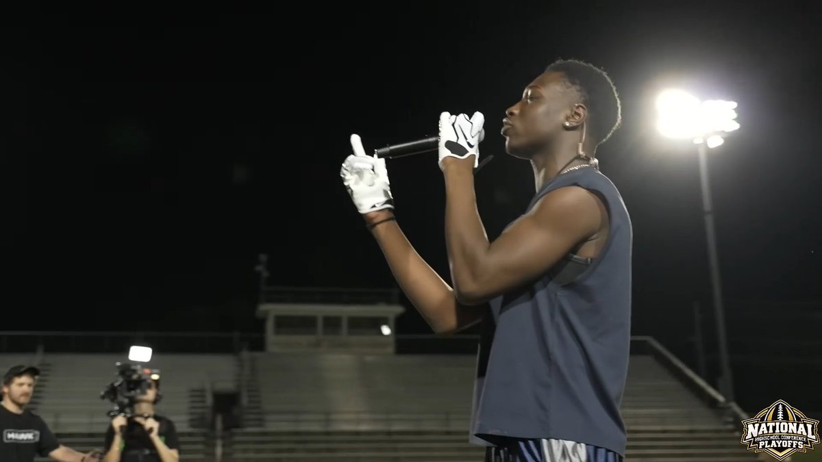 A young man performs on stage at night, holding a microphone and wearing white gloves, with a bright stadium light illuminating him from behind. The bottom right corner displays a "NATIONAL High School Conference PLAYOFFS" logo, while a camera crew films from the foreground.