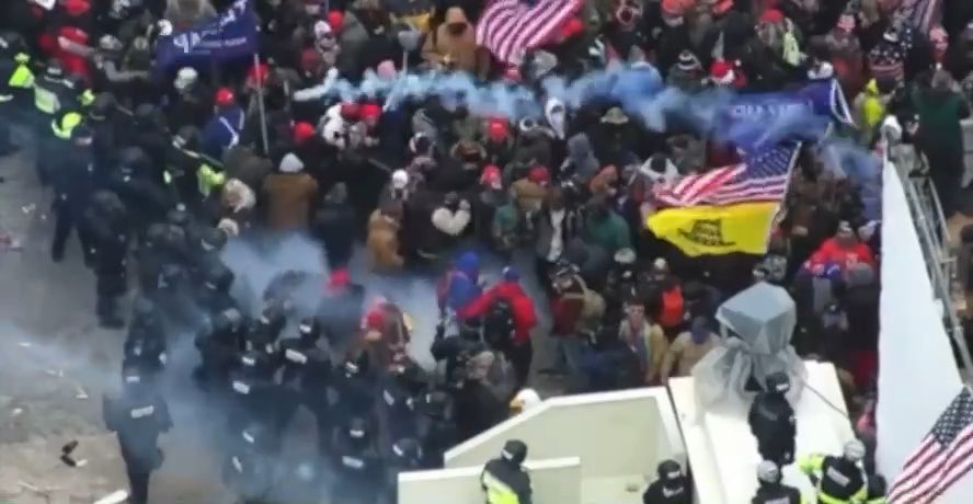 A chaotic scene from the January 6th Capitol breach shows police in riot gear forming a defensive line against a crowd of protesters, with smoke from tear gas billowing through the air and American flags and Trump campaign banners visible among the demonstrators. The image captures the moment referenced in the caption, illustrating the tense confrontation where crowd-control munitions were deployed amid the escalating clash between rioters and law enforcement.