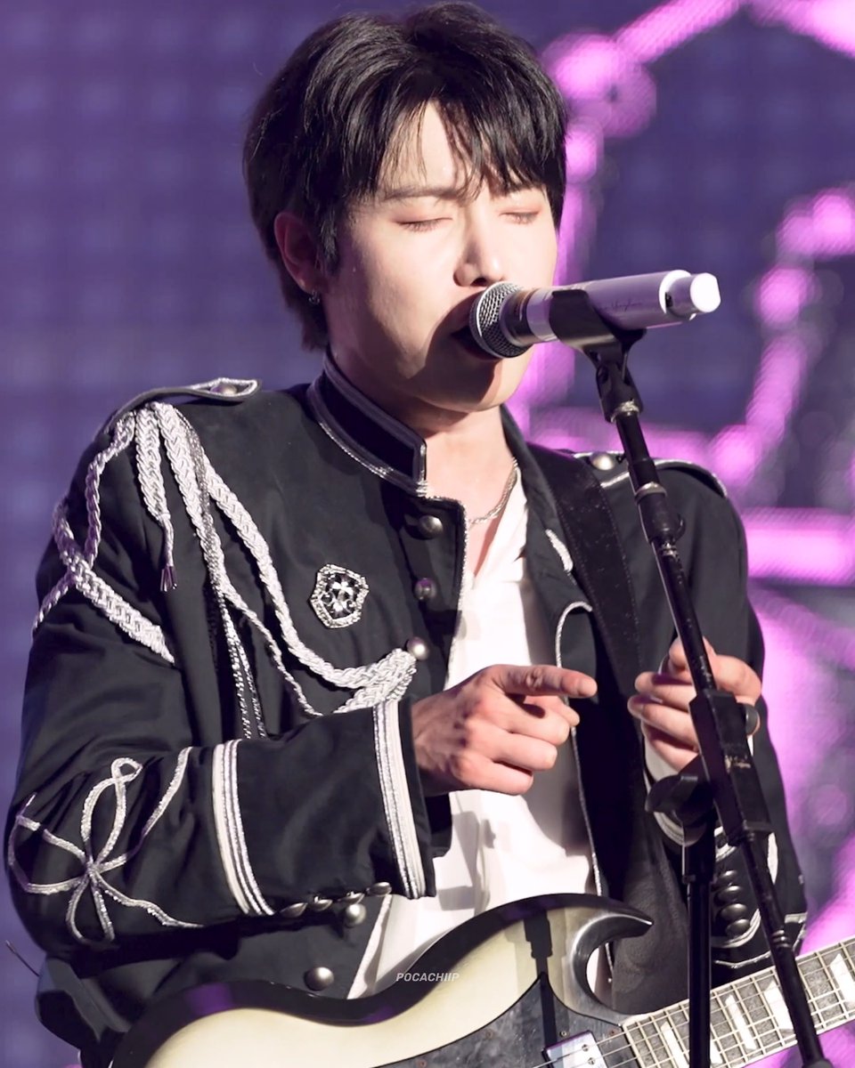 A young male performer sings with his eyes closed while playing a guitar, dressed in an ornate black military-style jacket with silver braided cords against a purple bokeh background.