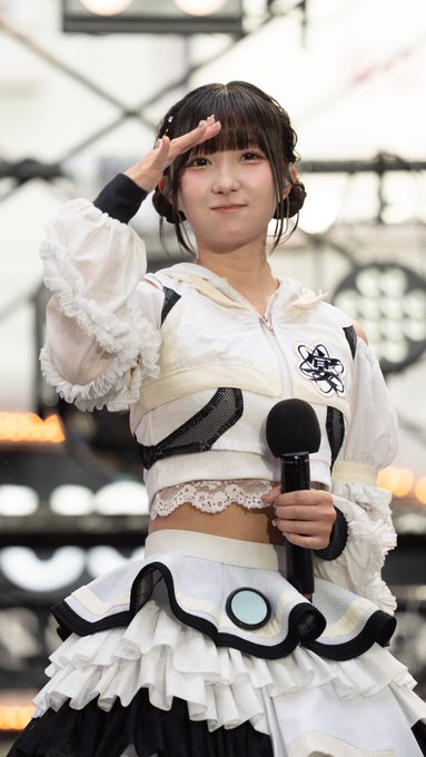 A young female idol in a white ruffled stage outfit with an atomic symbol performs a salute while holding a microphone.