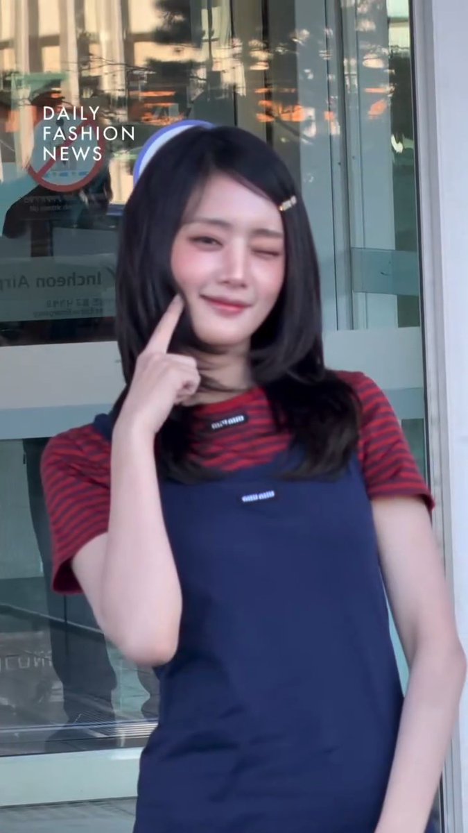 A young Asian woman wearing a red striped shirt under a navy blue pinafore dress winks at the camera while pointing to her cheek, with a 'DAILY FASHION NEWS' overlay.