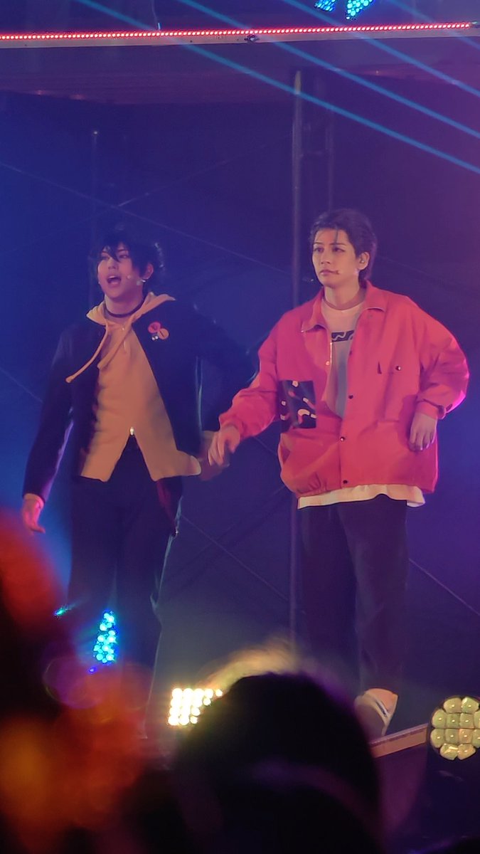 Two male performers hold hands on stage under blue and red lighting; one wears a yellow hoodie under a dark jacket, the other a pink jacket.