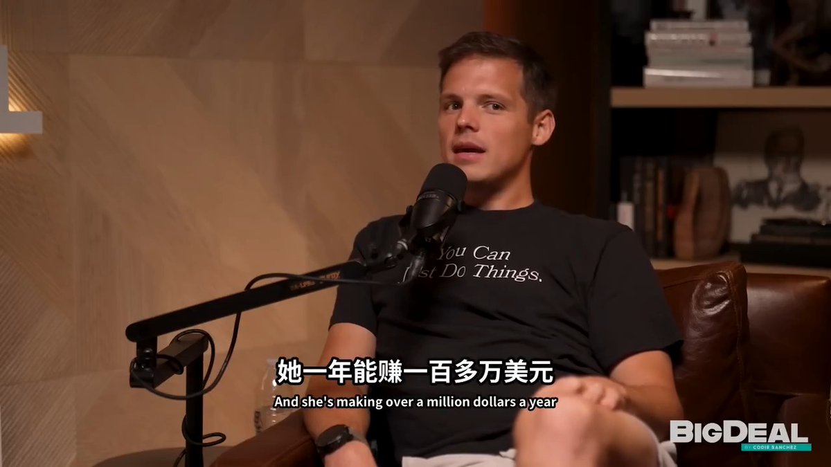 A man in a black t-shirt reading 'You Can Just Do Things' speaks into a microphone on the BigDeal podcast by Eddie Zantye, with subtitles translating his speech: 'And she's making over a million dollars a year.'