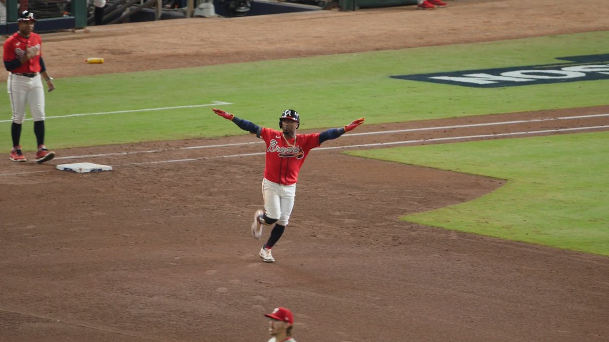 An Atlanta Braves player wearing a red "Braves" jersey celebrates with arms outstretched while rounding the bases, likely after hitting a home run that extended the team's lead. A coach in matching uniform stands near the dugout, and a large "WS" logo is visible on the field, hinting at the World Series setting.