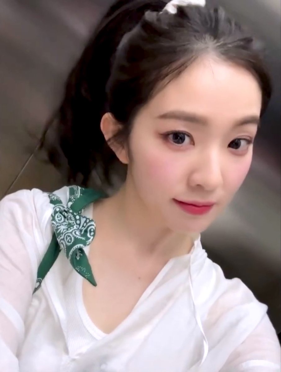 A close-up selfie of a young woman with her hair tied back, wearing a white shirt and a green bandana scarf around her neck.