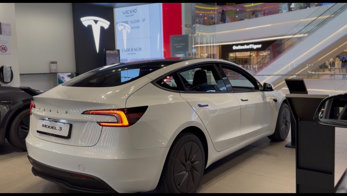 A white Tesla Model 3 displayed in a shopping mall showroom, with the Tesla logo visible in the background along with other retail brand signs.
