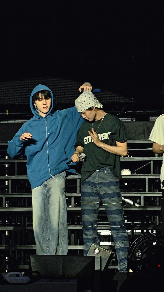 Two performers on a concert stage, one in a blue hoodie and baggy jeans with his hand on the other performer's bandana-covered head, while the other wears a black t-shirt reading 'ETT...VERT' and striped jeans.