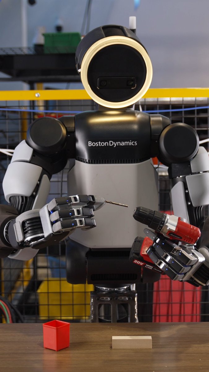 A Boston Dynamics humanoid robot holds a power drill and a drill bit, appearing to work on a small task with a red box and wooden block on the table in front of it.