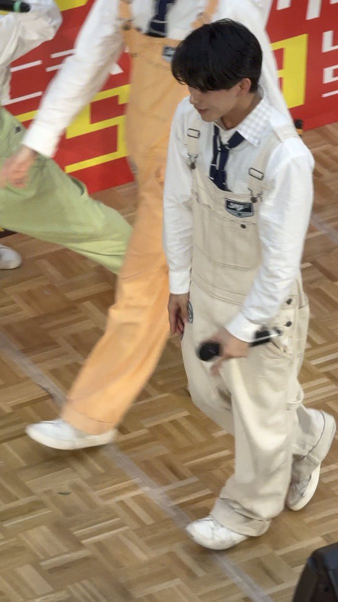A young man with dark hair wearing beige overalls and a white shirt, walking on a wooden floor while holding a microphone, appearing to be at an event or performance.