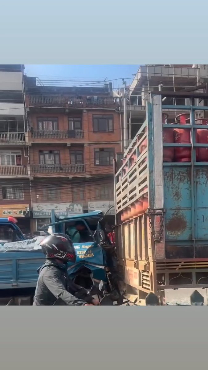 A street scene showing a motorcyclist next to a truck loaded with red gas cylinders, with old multi-story buildings in the background.