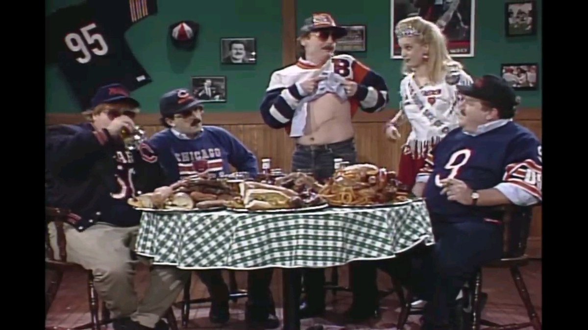 A scene from a comedy sketch shows five people wearing Chicago Bears jerseys, with one man lifting his shirt to expose his belly while a woman in a cheerleader outfit looks on, gathered around a table covered in a green and white checkered cloth with food on it.
