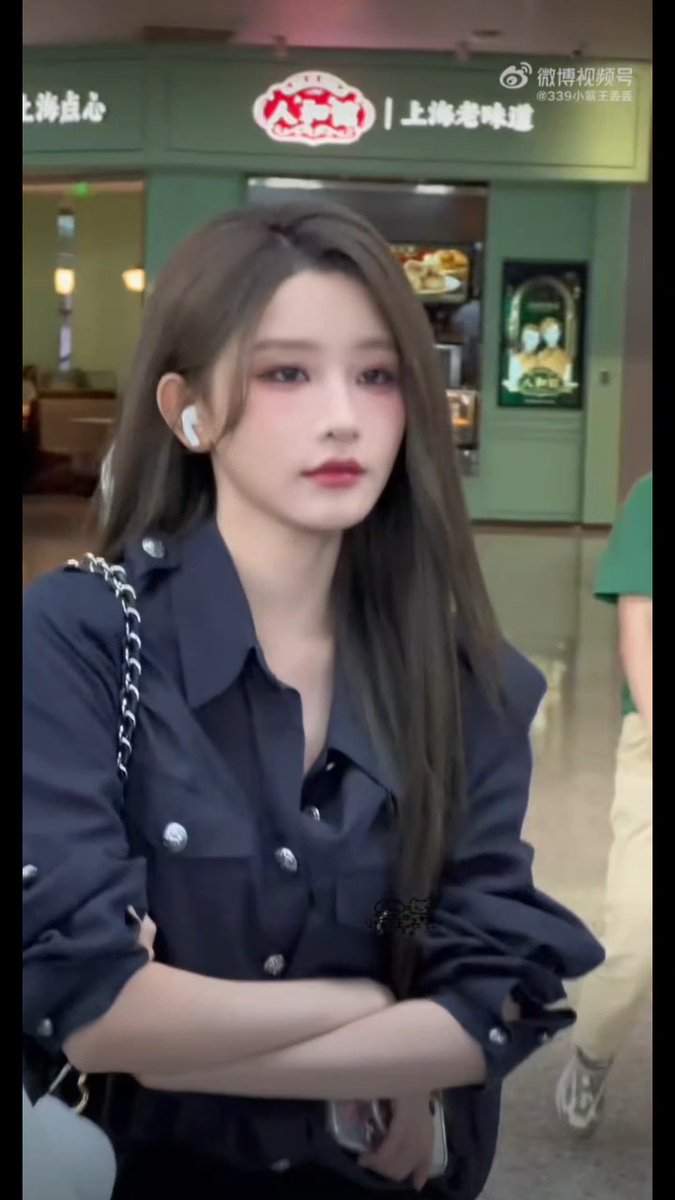 A young woman with long brown hair and an earbud stands with arms crossed in a navy blue button-up shirt in front of a green storefront with red Chinese characters.