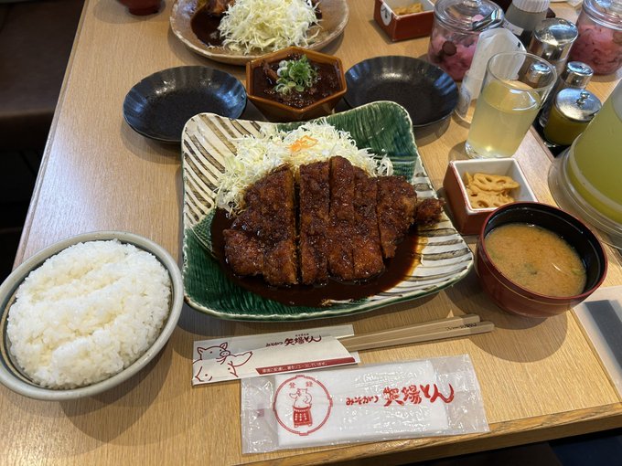A Japanese meal set featuring a breaded and fried cutlet covered in a dark sauce on a green plate, served with shredded cabbage, white rice, miso soup, and pickles on a tray reading 'I'm like 矢場とん'.