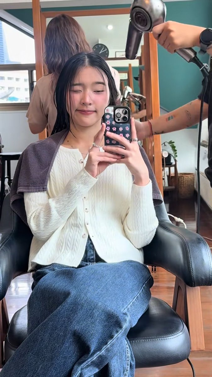 A young woman with dark hair sits in a salon chair wearing a white knit cardigan and jeans, taking a mirror selfie with a floral-cased phone while a stylist uses a hairdryer on her hair.