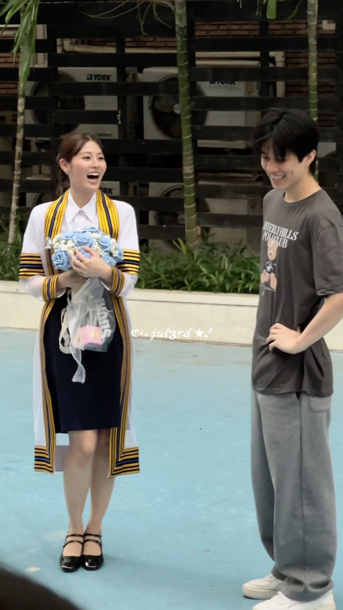 A young woman in a white graduation gown with yellow and black striped trim laughs joyfully while holding a bouquet of blue roses, standing next to a smiling young man in a gray "Beverly Hills Polo Club" t-shirt and gray pants. The watermark "juj3rd" appears on the image, and the setting appears to be an outdoor venue with greenery in the background, consistent with the graduation day celebration referenced in the post for Pahnthitta and Aston.