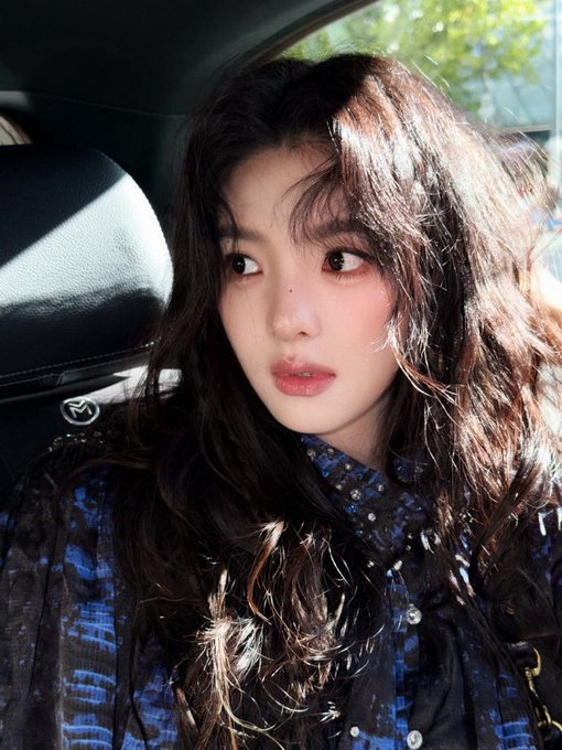 A young woman with long wavy brown hair and glossy lips sits inside a car taking a selfie, wearing a dark blue patterned top.