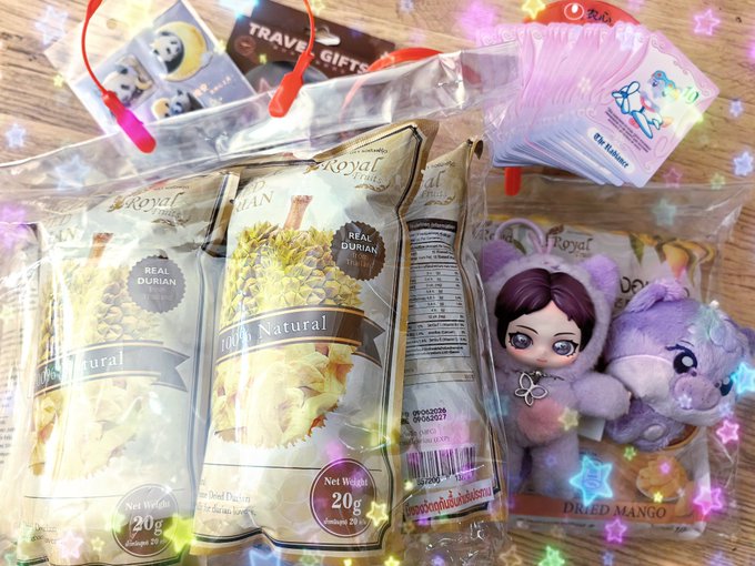 An assortment of Thai dried fruit snacks displayed on a wooden surface, including 'Royal' brand durian chips and 'Dried Mango,' along with cute character-themed packaging, decorated with sparkle filter effects.
